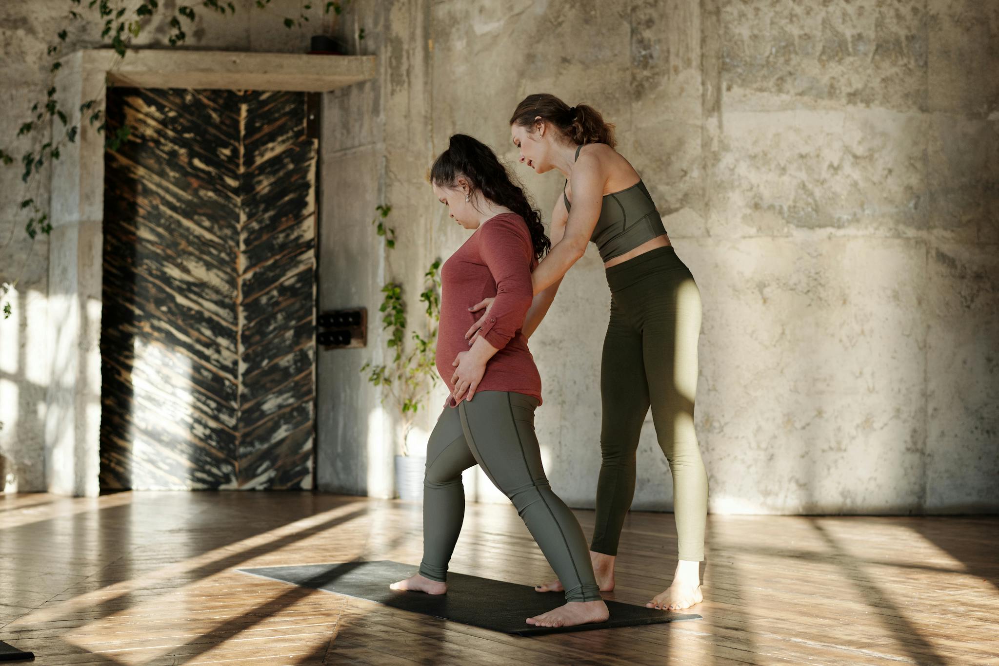 A woman supports another woman with Down syndrome during a yoga session indoors.